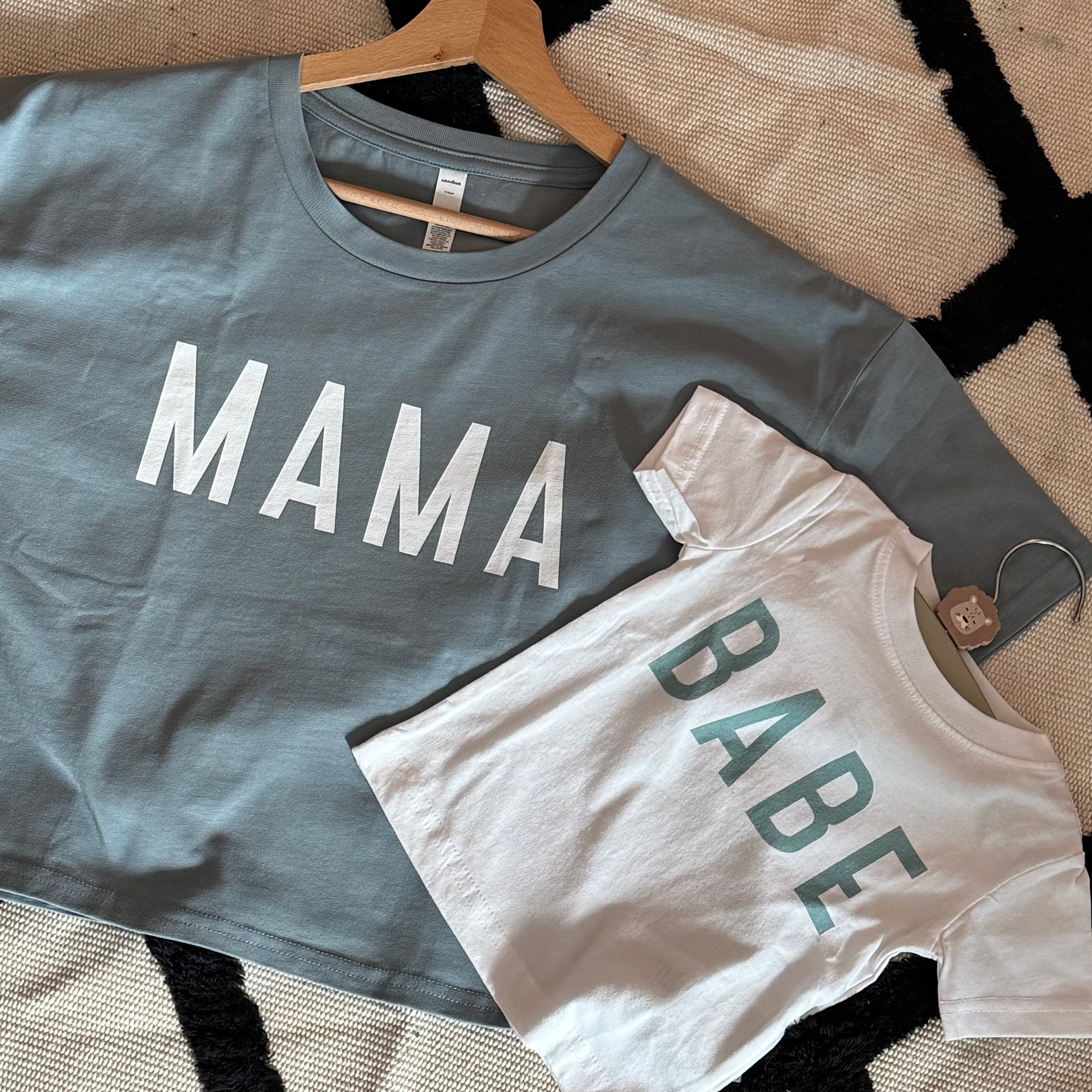 Blue 'MAMA' t-shirt and white 'BABE' onesie on a textured surface