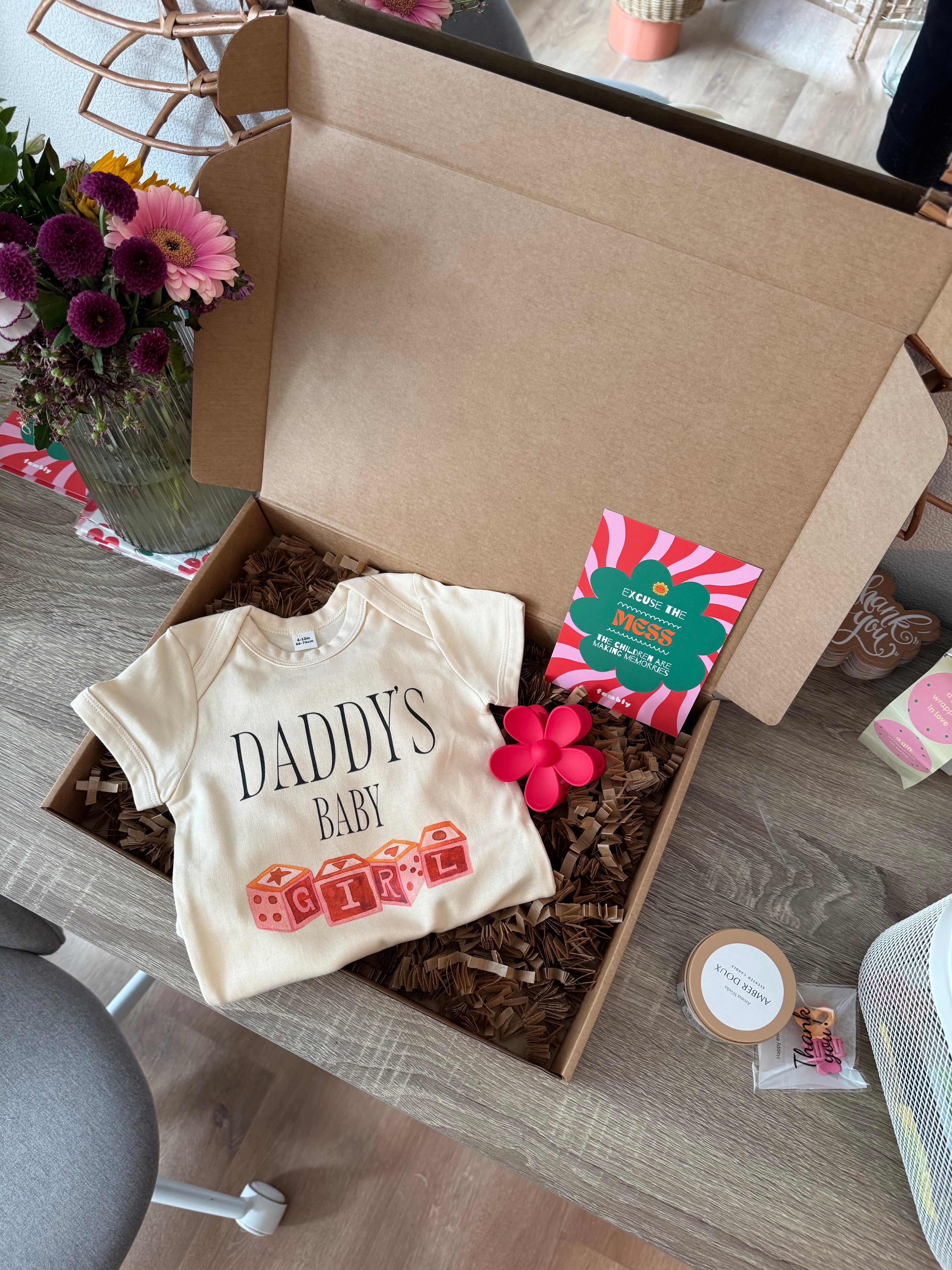 Open box containing baby clothes with a 'DADDY'S BABY' shirt on a wooden floor.
